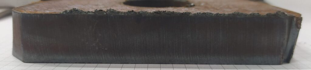 Close-up of a metal workpiece cross-section after cutting, showing a rough upper edge and a darker heat-affected zone along the cut surface.