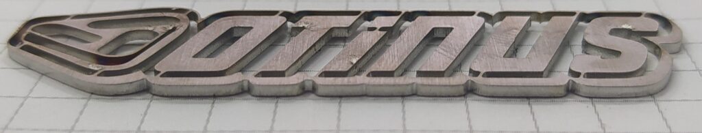 Laser-cut metal OTINUS logo with smooth contours and visible heat marks along the edges, highlighting precise cutting quality. The layered steel finish emphasizes industrial craftsmanship and brand identity.