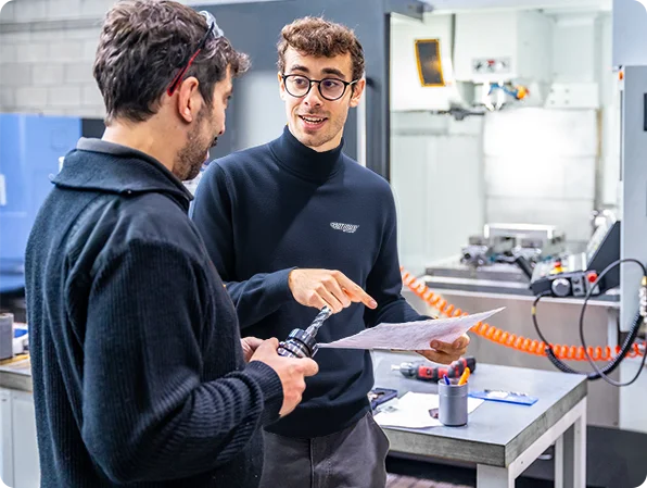Two engineers discussing technical documentation in a manufacturing workshop, reviewing a component and production details.