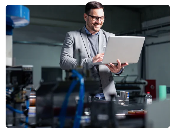 Smiling engineer using a laptop to monitor production processes in a modern industrial workshop.