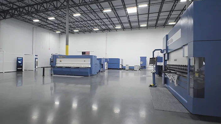 Spacious industrial showroom with Otinus sheet metal machines, including press brakes and CNC systems, arranged on a polished concrete floor under a high, well-lit ceiling.