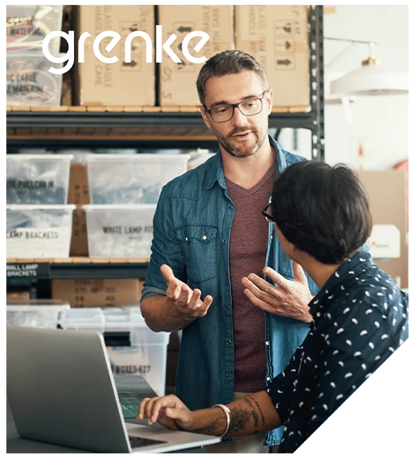 Dwóch mężczyzn prowadzi rozmowę w magazynie przy otwartym laptopie; jeden gestykuluje, drugi siedzi przy komputerze, a w tle stoją pudełka i regały.