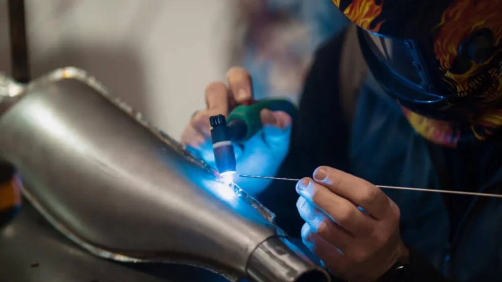 Spawacz w masce ochronnej wykonujący precyzyjne spawanie TIG elementu ze stali nierdzewnej, prowadząc drut spawalniczy w świetle niebieskiego łuku.