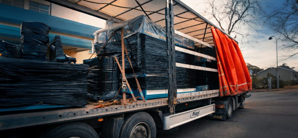 Załadunek maszyn przemysłowych na naczepę ciężarówki, zabezpieczonych folią i pasami transportowymi, przygotowanych do bezpiecznego przewozu w ramach logistyki przemysłowej.