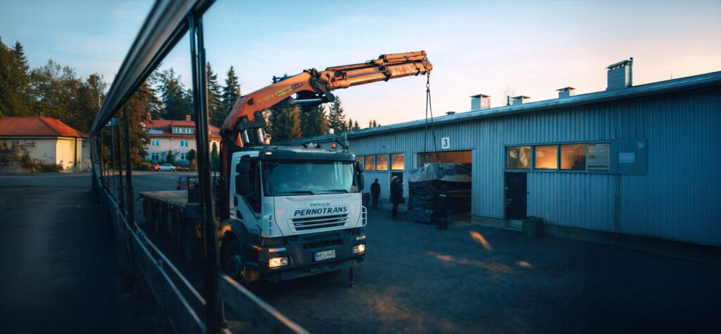 Załadunek maszyny przemysłowej przy hali produkcyjnej z użyciem ciężarówki z żurawiem hydraulicznym, realizowany w zorganizowanym procesie logistyki przemysłowej.