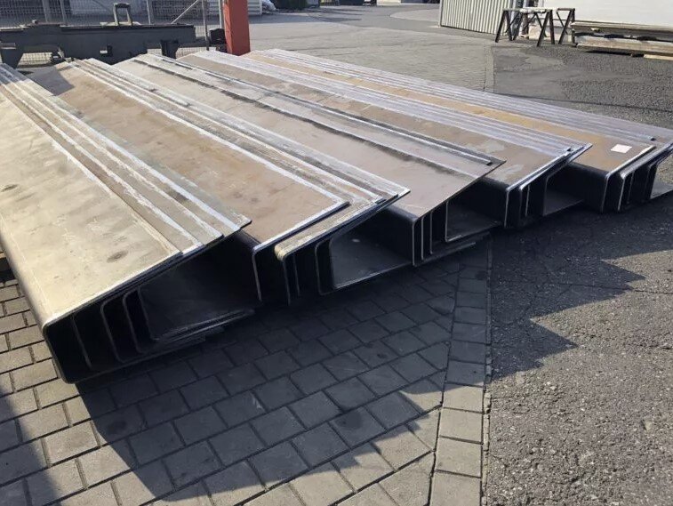 Stacked galvanized steel U-profiles laid flat on an outdoor paved surface. The profiles are uniform in shape and size, prepared for transport or further industrial processing.