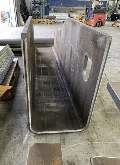 Large bent steel U-channel with smooth radiused corners, standing upright in an industrial workshop. The image shows thick sheet metal formed into a deep profile with cut-out openings on the side walls.